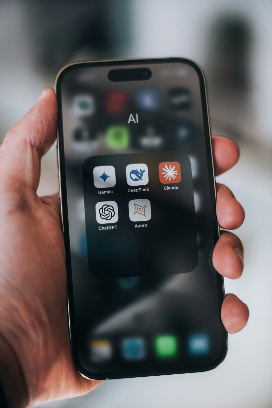 A hand holding a smartphone showing a folder of AI apps including Gemini, DeepSeek, Claude, and ChatGPT